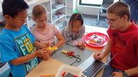 Four young students are sitting around a classroom table building a contraption out of legos and wires. It looks like it might be able to move prompted by technology.