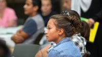 A teenage girl is sitting at a table among other students sitting at tables in a classroom. Everyone else is blurred out.