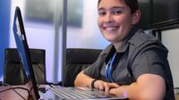 A young boy is sitting at a desk with a laptop in front of him. He is smiling directly at the camera. A TV is on the wall behind him, and two leather chairs are to the right of him against a clear wall, adjacent to a similar room with a TV on the wall.