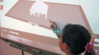 A closeup of a young girl standing next to and pointing at a projector screen. A 3D drawing made up of small squares is projected on the screen, showing the inside of a white room with a brown floor and a small white pyramid in the middle.