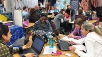 Three groups of four to five teachers are sitting at tables in a classroom talking to each other, looking at their laptops, and writing on paper.