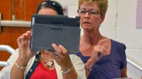 Two adult women are standing in a classroom with tablets in their hands. One of them is holding up their tablet, and both women are looking at it.