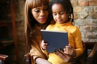 A daughter sitting on her mother's lap, both looking at a tablet that the daughter is holding