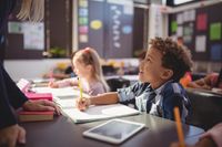A young boy sitting at his desk, looking up at his teacher who is talking to him, and smiling