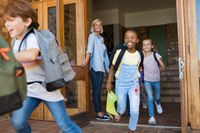 A teacher letting her students out the front door of the school on the last day of the school year. The kids are running with smiles on their faces, and the teacher is smiling as she watches them go.