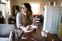A woman sitting at her kitchen table with a cup of tea, writing in a notebook and looking thoughtfully out the window