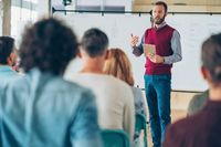 A teacher gesturing in front of a whiteboard to a room of his fellow teachers and faculty