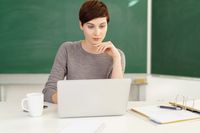 A teacher using her laptop in a classroom. She's sitting in front of a blackboard and has a notebook next to her.