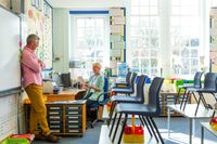 A teacher and a principal meeting in a classroom after school. The teacher is sitting at her desk and the principal is standing, leaning against the whiteboard.