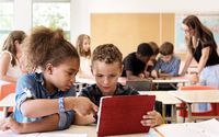 A classroom full of kids using tablets. The teacher is helping a group in the background and two students are working together in the foreground.