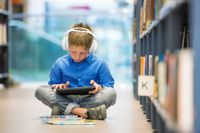 An elementary school student sitting on the floor of a library with a tablet and headphones, listening to an audiobook