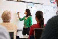 A teacher standing in front of a classroom full of her colleagues. She's writing something on a whiteboard.