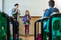An elementary student presents her findings for a PBL project to her class. Her teacher stands next to her, applauding.