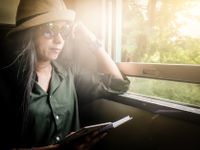 A woman in sunglasses and a hat sitting on a train on a sunny day with the window cracked open, reading a book