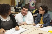 High School teacher kneeling down to talk to 2 students sitting at their desks