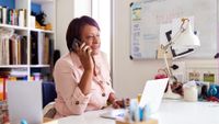 Woman at home speaking on phone sitting at desk with laptop