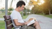 Man reading a book on park bench with his dog sitting next to him