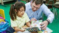 Teacher kneeling down and reading with his elementary student at her desk