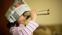 Young girl playing make believe with a newspaper hat and looking through paper roll. 
