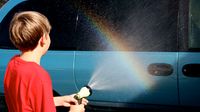 Boy sprays water from garden hose onto car and makes a rainbow.