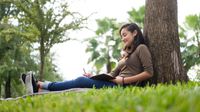 Girl writing in her notebook while sitting against a tree outside