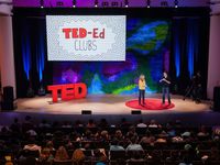 A male and female adult are standing on a stage with a large audience in front of them. Standing on the stage are three big, red letters spelling, "TED." On a screen behind them says, "Ted-ed clubs."