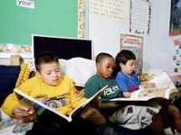Three young boys are sitting on a couch in their classroom, reading.