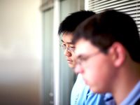 A closeup of two young, adult males looking dejected with their heads down. They're standing against a window with closed blinds.