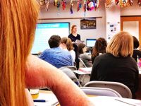High school students are sitting at desks in foldable chairs. A teacher is standing at the front of the class next to a large screen on the white board. The screen is displaying a presentation slide that says, "Language Acquisition."