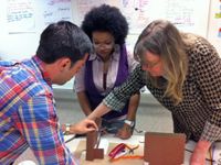 One male and two female teachers are leaning over a classroom table, building something together. They have two pieces of cardboard cut, standing up like mini buildings, one with an open door, and a piece of wire is connecting them.