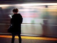 A woman in a coat is standing by a moving subway, reading a book.