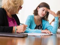 A short-haired, blond female teacher in a pink shirt and black blouse is sitting next to a brunette teenager girl, helping her study.