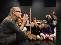 A female teacher is sitting cross-legged on a stage, and students are standing, talking to each nearby in costumes.