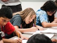 Students are hunched over their desks, writing in notebooks, with their teacher looking upon them.