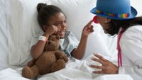 A young girl is lying in a hospital bed, holding a teddy bear, smiling, and pointing at her doctor's face. Her doctor is wearing a clown hat and nose.