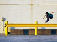 A young teenage boy in a teal t-shirt and brown pants is in the air on a skateboard. Both of his arms are extended out to his sides and his knees are up to his chest. He's hovering over a yellow beam against an off-white brick wall. 
