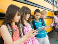 Five young students are leaning against a bus all looking at mobile devices.