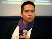 An adult male is sitting, holding a microphone, and speaking to someone. He's wearing glasses perched on top of his head, a white top, black coat, purple scarf, and a conference name tag on his chest. 