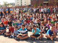A lot of young students are sitting outside on a blacktop, with teachers standing behind them, all facing the same direction.