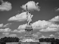 A black and white photo of the statue of liberty -- a woman in a toga, a crown, and her right arm raised up in the air, holding a torch. Behind her, the sky is filled with clouds.