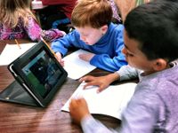 A group of elementary students are sitting at circular tables, sharing a tablet, and working on a paper assignment.