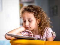 A young girl with curly, light brown, golden hair and a pink shirt is sitting, resting her arm on a yellow couch, reading on a tablet in her house.