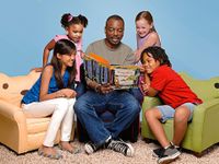 LeVar Burton, associated with Reading Rainbow, is sitting on a blue couch chair reading a book to four kids. A girl is sitting on a cat chair to his left, a boy is sitting on a frog chair to his right, and two girls are standing behind them.