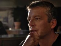 A closeup, neck up, of a man in a dark room, staring off in thought with his hand in a fist against his chin.