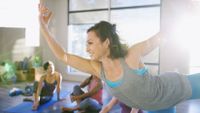 A smiling woman in a yoga class standing on one leg and both arms are extended.