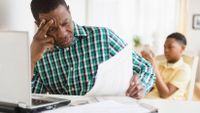 A dad is sitting at the kitchen table with a laptop in front of him, looking at paper in his hand in frustration. His son is sitting behind him, looking at his phone.