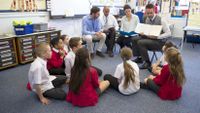 Four adults sit in front of a circle of students—one adult instructs and the other three observe.