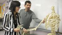 Girl holding the lower arm of a skeleton while her teacher points out something near the shoulder