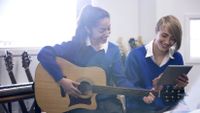 One girl in a school uniform plays guitar while looking at a tablet computer that a girl next to her is holding.