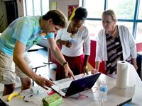 Three teachers are standing by a desk in a classroom, working on a project connected to a computer.